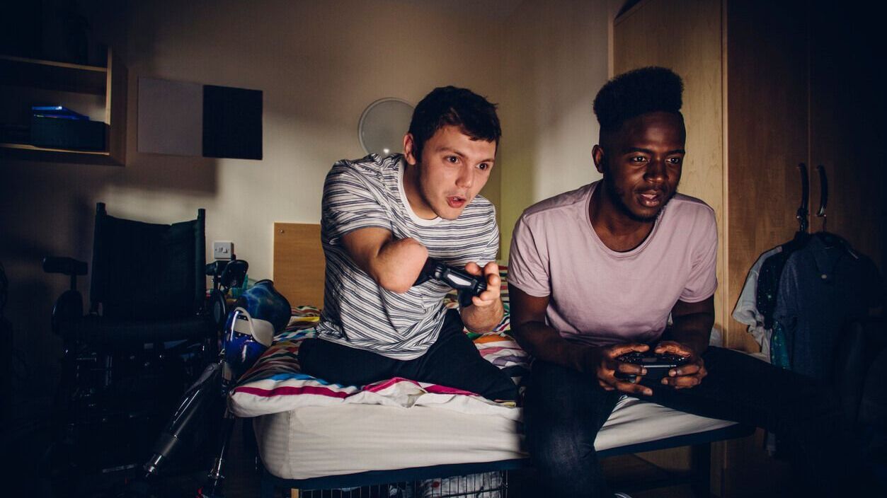 Inklusives Wohnen: Ein junger Mann mit Handicap sitzt zusammen mit einem anderen jungen Mann auf dem Bett und spielt mit der Spielekonsole