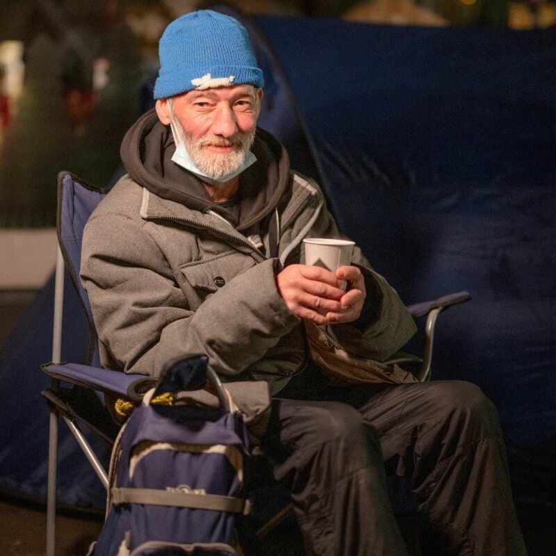 Ein älterer Mann mit blauer Mütze sitzt mit einem Pappbecher in der Hand auf einem Campingstuhl