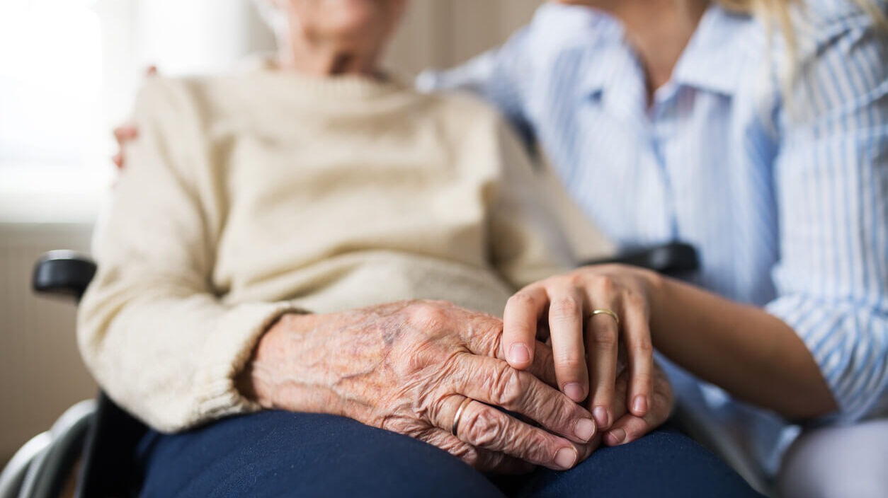 Eine ambulante Pflegekraft hält die Hand einer Seniorin im Rollstuhl.