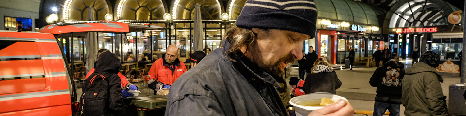 Ein bedürftig aussehende Mann steht vor einem Bahnhof und isst Suppe.