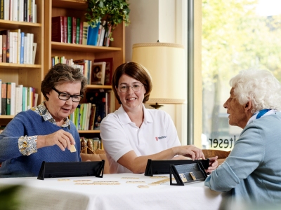 Malteser Mitarbeiterin spielt mit zwei älteren Damen an einem Tisch. 