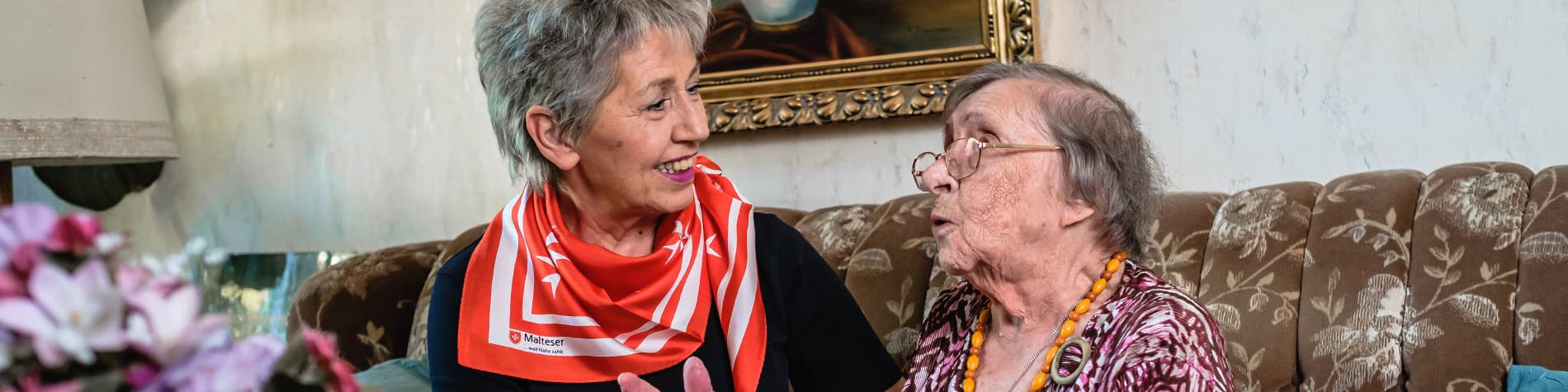 Malteser Mitarbeiterin spricht mit einer älteren Dame auf der Couch.