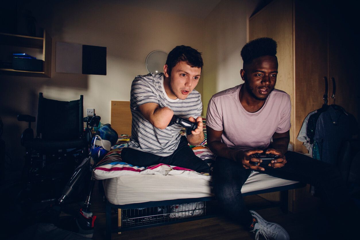 Inklusives Wohnen: Ein junger Mann mit Handicap sitzt zusammen mit einem anderen jungen Mann auf dem Bett und spielt mit der Spielekonsole