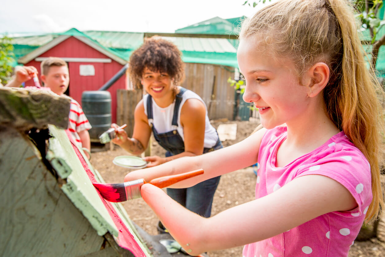 Junges Mädchen ohne Hände malt ein Holzhaus an in Begleitung mit einer Helferin