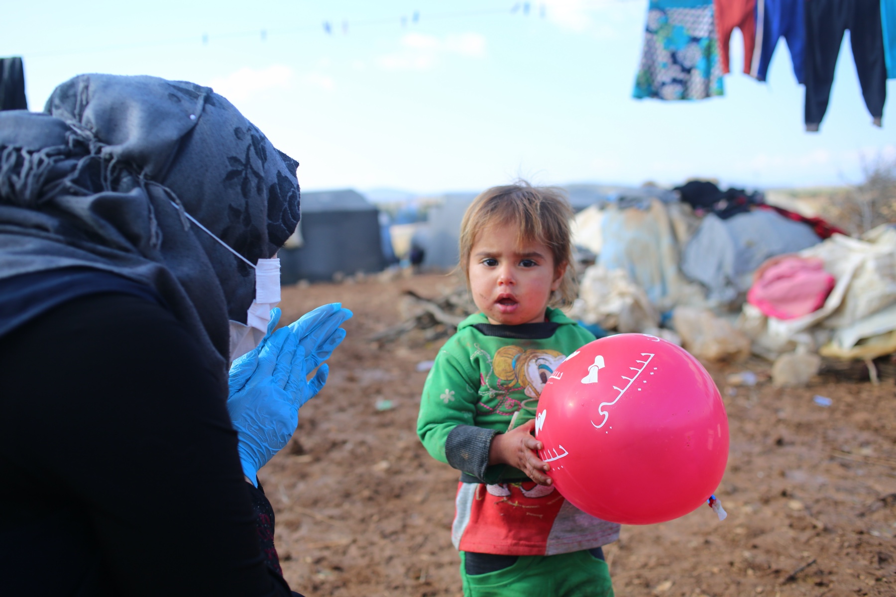 Kind mit einem Luftballon und eine Frau mit Kopftuch und Hygienehandschuhen in einem Flüchtlingscamp.