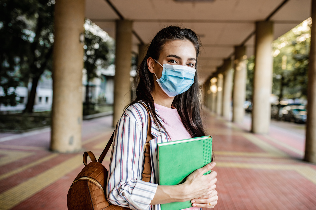 Junge Frau mit Hefter steht mit Mund-Nasen-Schutz auf einem Campus-Gelände.