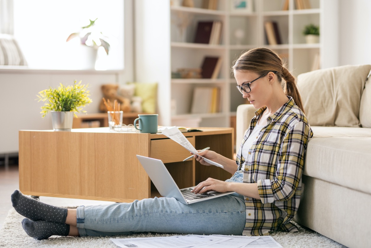 Junge Frau mit Brille sitzt auf dem Boden mit Laptop, Stift und einem Zettel.
