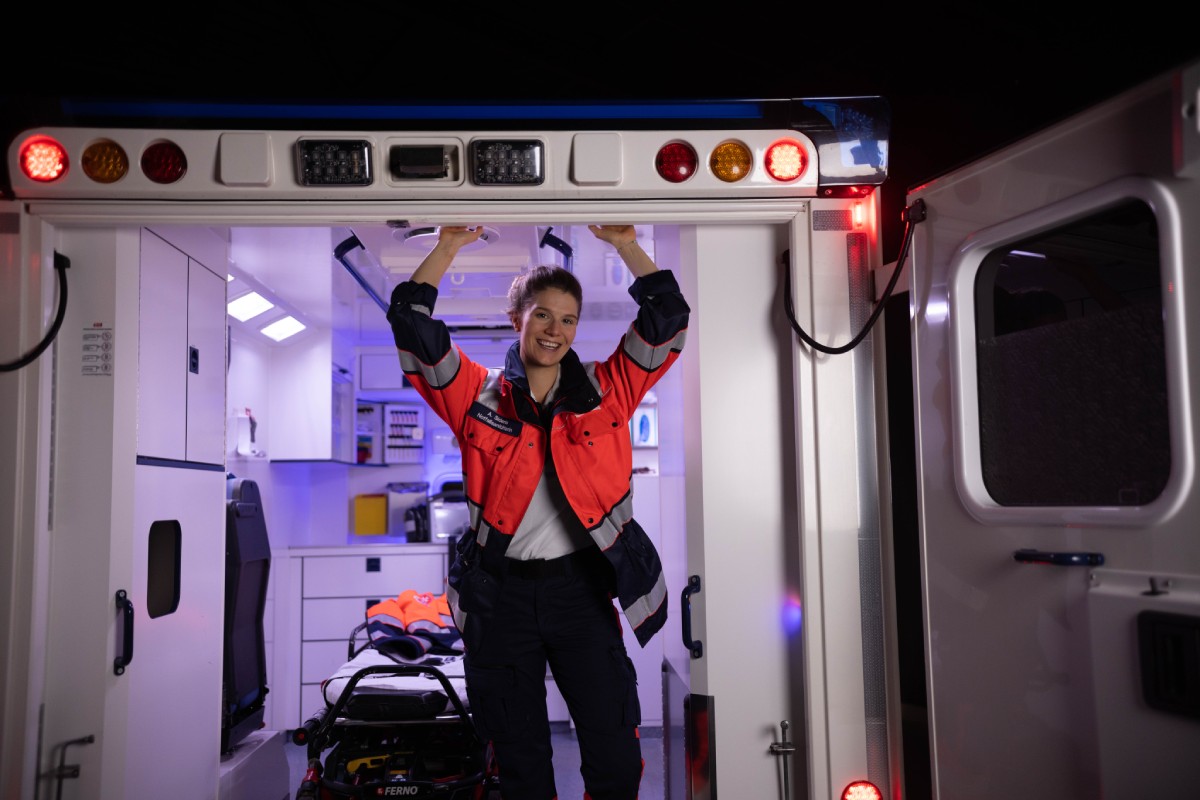 Eine junge, lächelnde Notfallsanitäterin im Heck eines Rettungswagens
