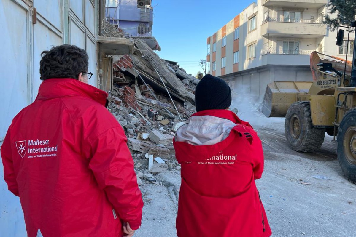 Zwei Menschen mit roten Jacken vor einem zerstörten Haus.