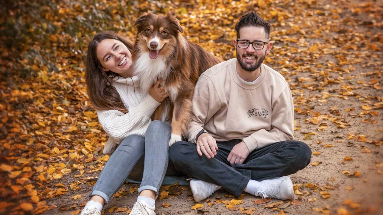 Eine junge Frau und ein junger Mann sitzen mit einem Hund auf Herbstlaub