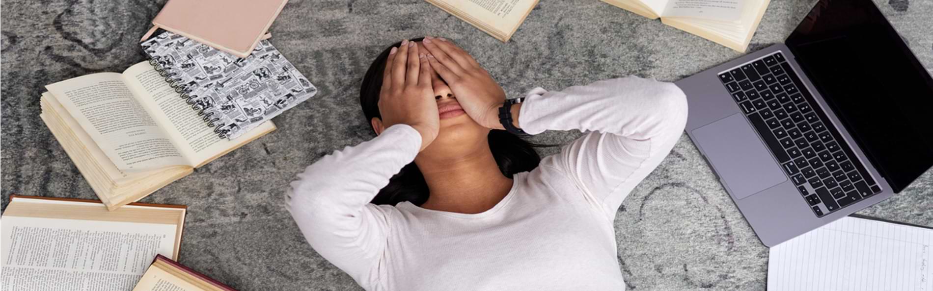Eine junge Frau liegt erschöpft zwischen Büchern, einem Laptop und einem Smartphone auf einem Teppich