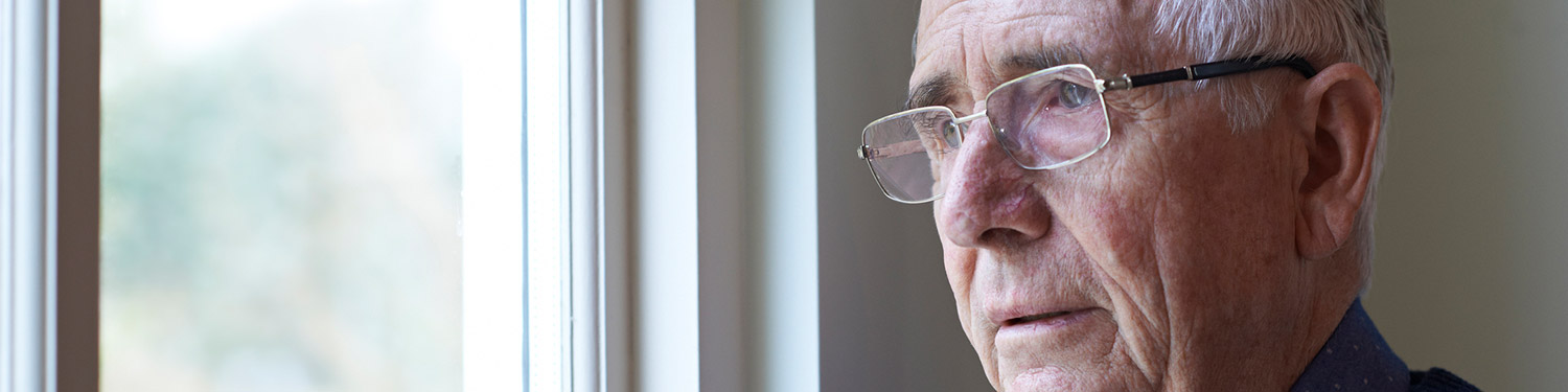Ein älterer Mann mit Brille und blauem Pullover schaut besorgt aus dem Fenster.