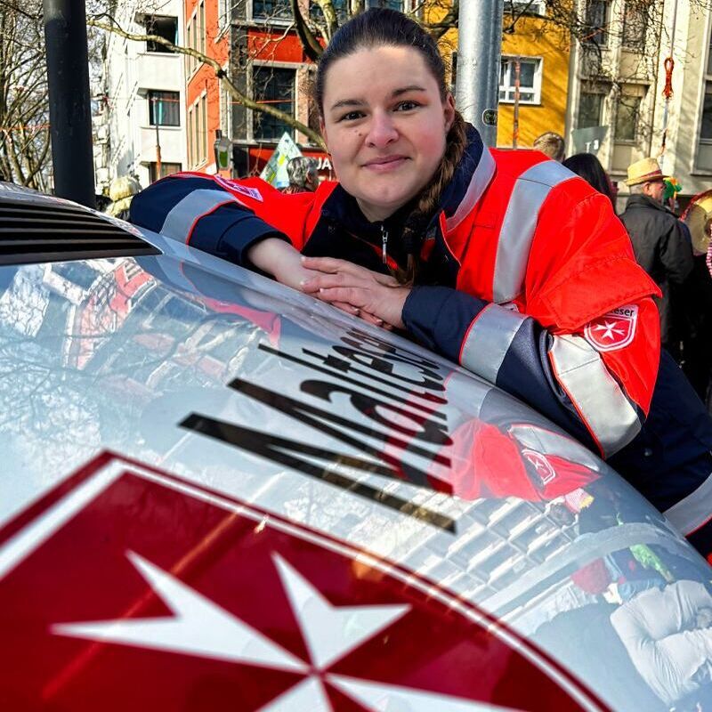 Eine Frau in Einsatzkleidung lehnt sich mit beiden Händen auf deine Motorhaube von einem Fahrzeug, auf dem „Malteser“ steht und das Wappen der Malteser zu sehen ist.