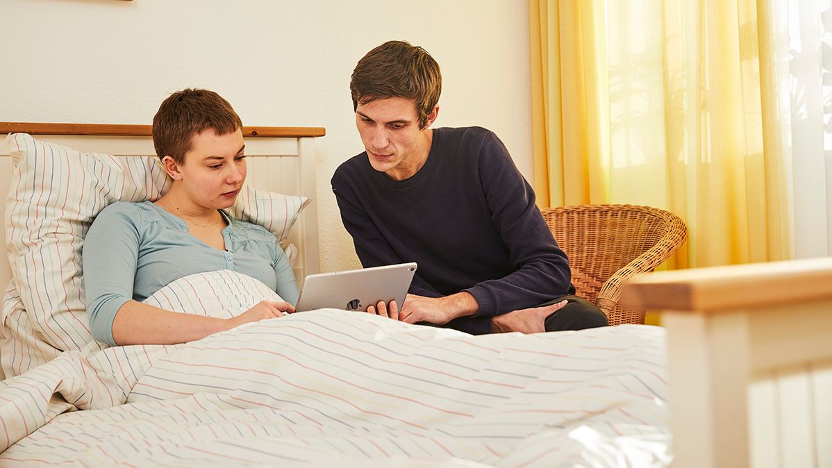 Junger Mann schaut sich auf einem Tablet etwas gemeinsam mit einer jungen Frau mit kurzen Haaren an, die in einem Bett liegt.