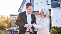 Eine ältere Frau zeige einem Mann den Hausnotrufknopf, den sie am Arm trägt.
