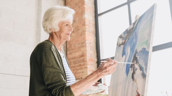 Ältere Frau steht vor einem Fenster und malt ein Bild.