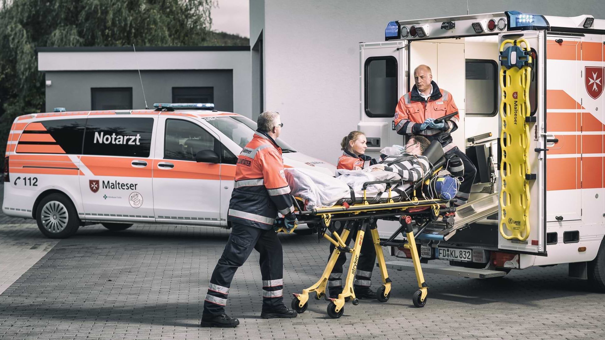 Malteser versorgen einen Patienten mit Beatmungsgerät auf einer Trage vor einem Notarzt- und Rettungswagen der Malteser.