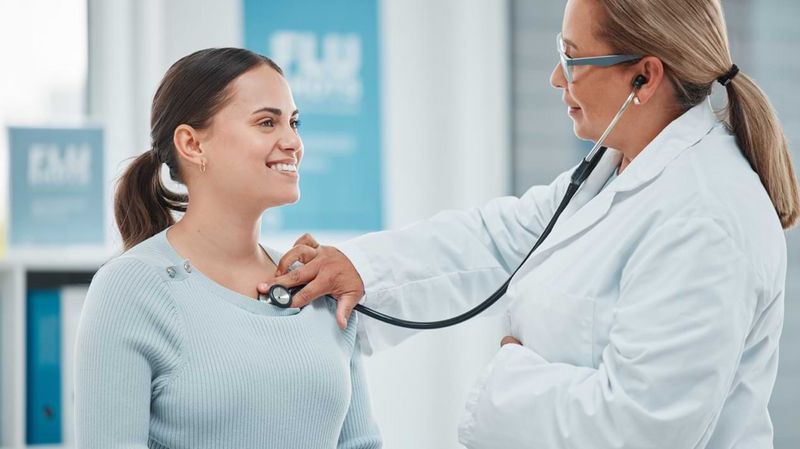 Eine junge Frau wird von einer Ärztin mit einem Stethoskop abgehört.
