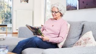 Eine ältere Dame sitzt auf einem Sofa und liest eine Zeitschrift.