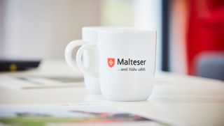 Weisse Malteser Tasse bedruckt mit Malteser Logo und Malteser–weil–Nähe–zählt-Schriftzug.