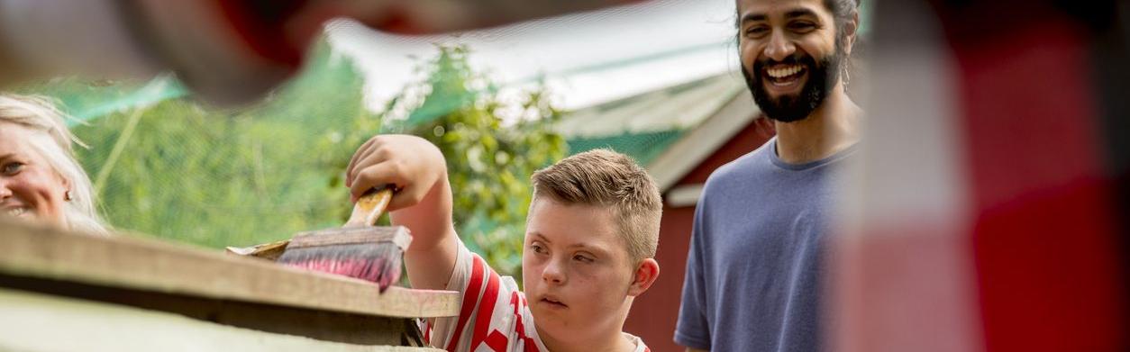 Inklusion in Deutschland: Junge streicht mit einem Betreuer eine Holzhütte an