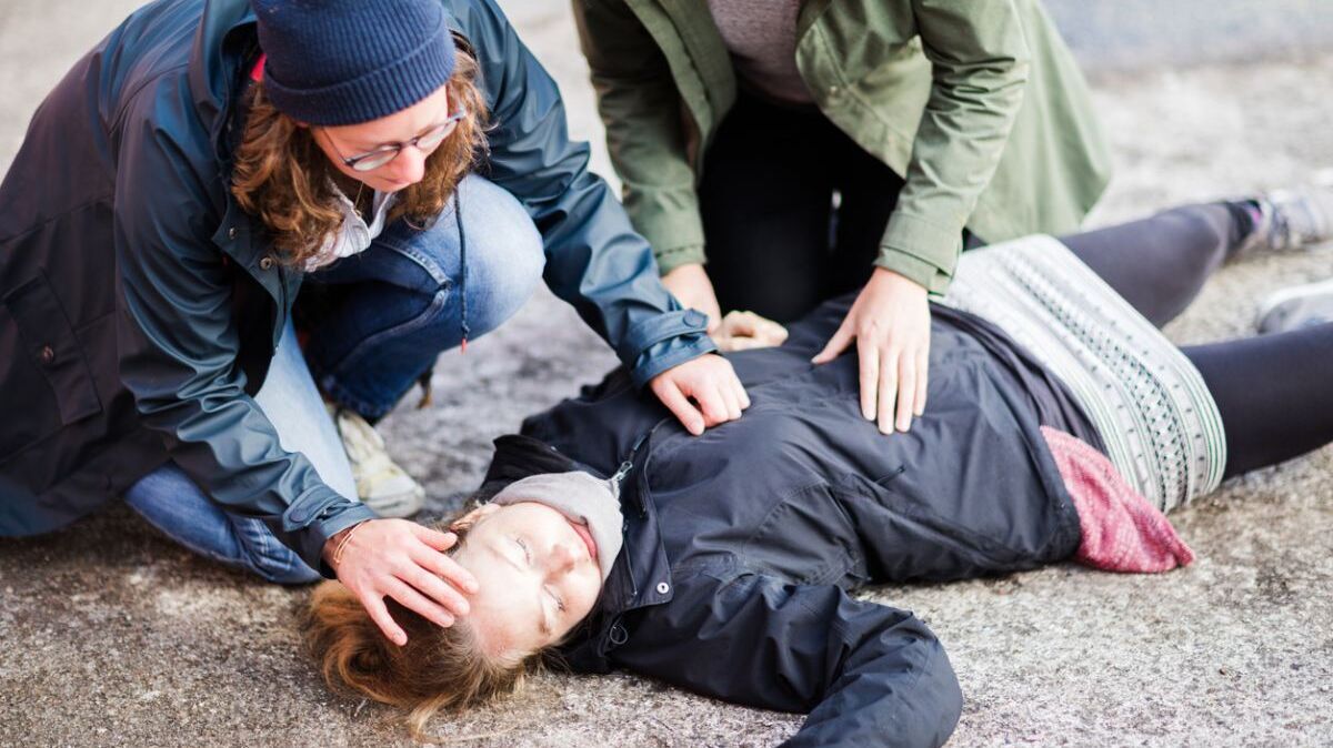 Zwei Frauen knieen auf einer Asphaltstraße über einer bewusstlosen Frau.