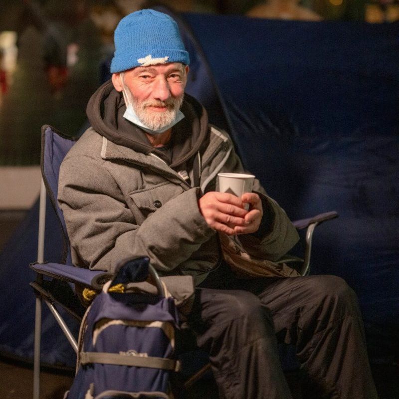 Ein Mann mit einer blauen Mütze sitzt auf einem Campingstuhl und trinkt aus einem Pappbecher