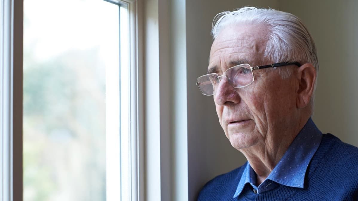Ein älterer Mann mit Brille und blauem Pullover schaut besorgt aus dem Fenster.