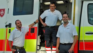Drei Mitarbeiter des Malteser Rettungsdienstes stehen in Einsatzkleidung am Rettungswagen.