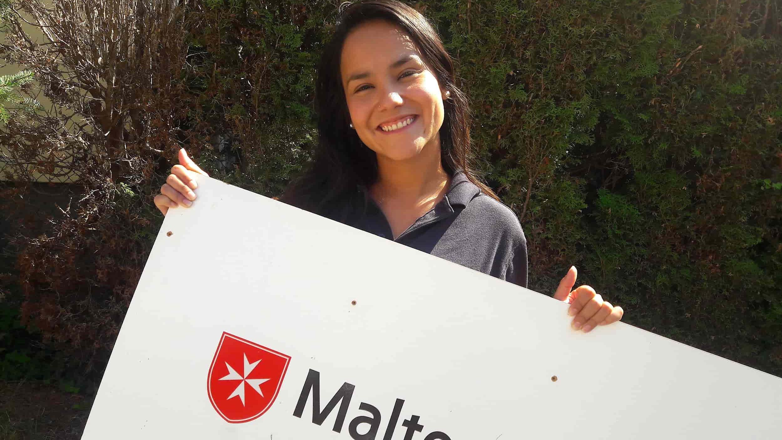 Junge, lächelnde Frau hält ein Schild mit Malteser-Logo