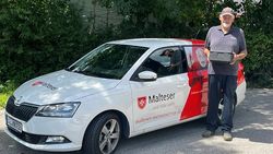 Ein Malteser Mitarbeiter steht mit einer Isolierbox in der Hand neben einem Malteser Auto.