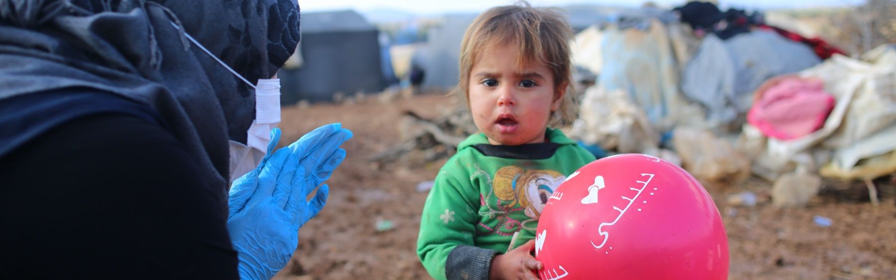 Kind mit einem Luftballon und eine Frau mit Kopftuch und Hygienehandschuhen in einem Flüchtlingscamp.