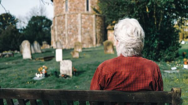 Älterer Mann sitzt auf einer Bank auf einem Friedhof.
