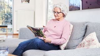 Ältere Dame mit Hausnotruf-Knopf an einer Kette liest auf der Couch eine Zeitschrift.