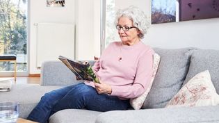 Ältere Dame mit Hausnotruf-Knopf an einer Kette liest auf der Couch eine Zeitschrift.