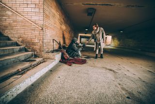 Unter einer Unterführung begegnet eine Frau einem dort sitzenden Obdachlosen und streckt ihm ihre Hand entgegen. 