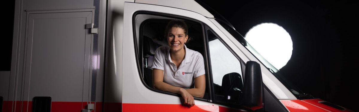 Eine junge Notfallsanitäterin schaut lächelnd aus dem Beifahrerfenster eines Rettungswagens.