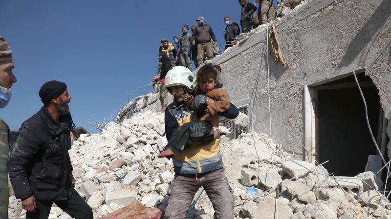 Ein Mann mit Helm trägt ein Kind über ein Trümmerfeld