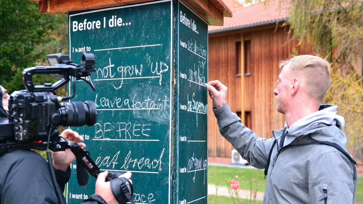 Junger Mann schreibt etwas mit Kreide an eine Tafel und wird dabei gefilmt