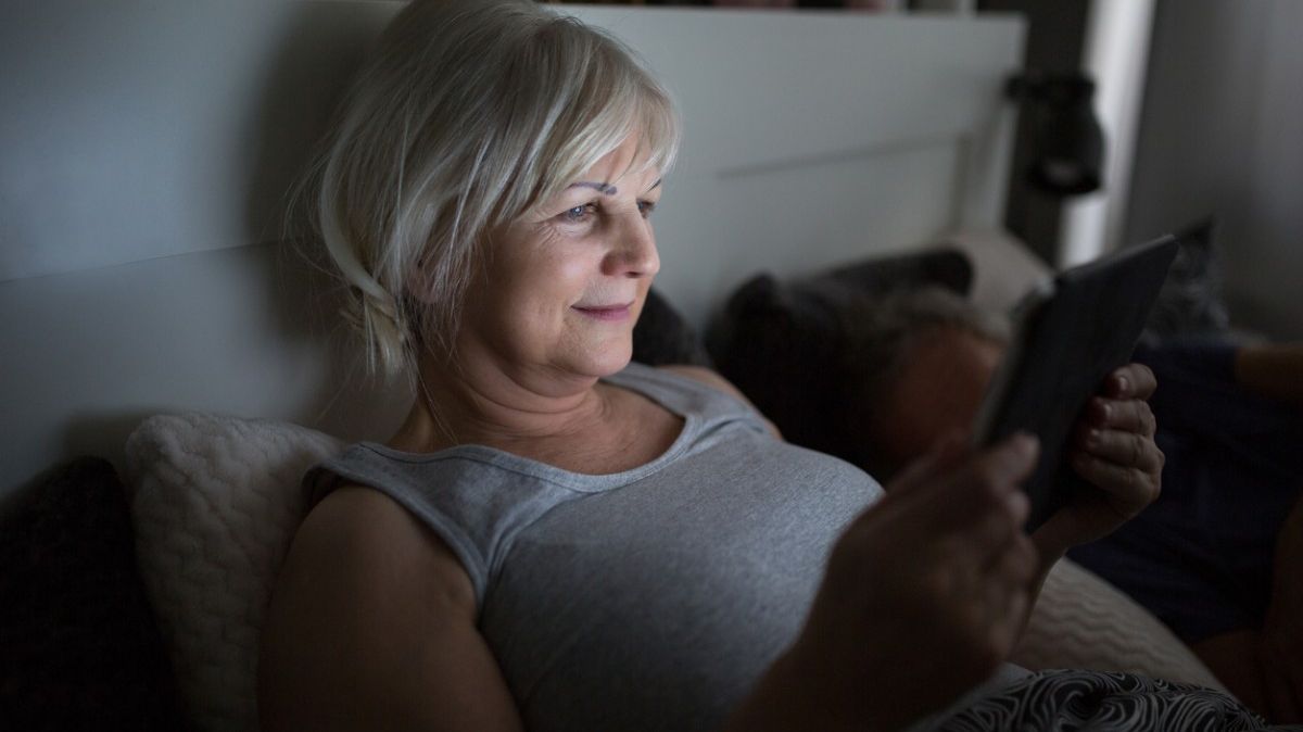 Ältere, lächelnde Frau liest im Bett mit einem E-Book-Reader.