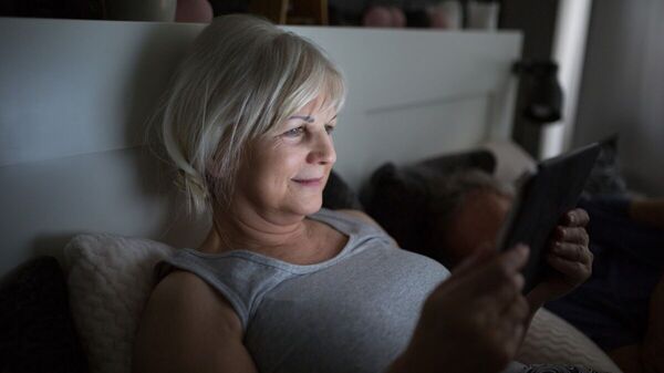 Ältere, lächelnde Frau liest im Bett mit einem E-Book-Reader.