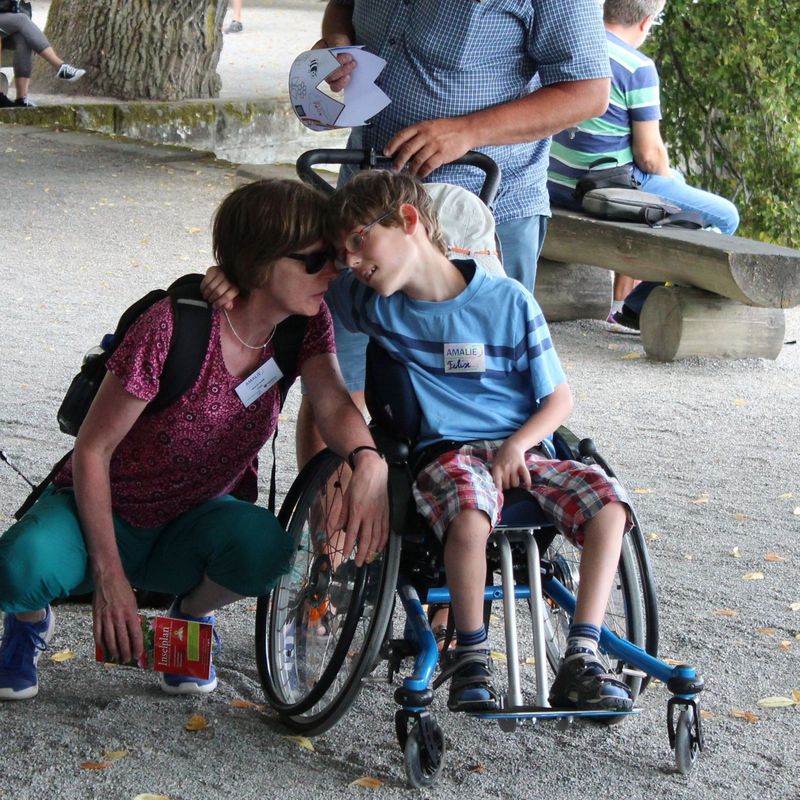 Eine Frau mit Sonnenbrille kniet bei einem Jungen, der in einem Rollstuhl sitzt. 