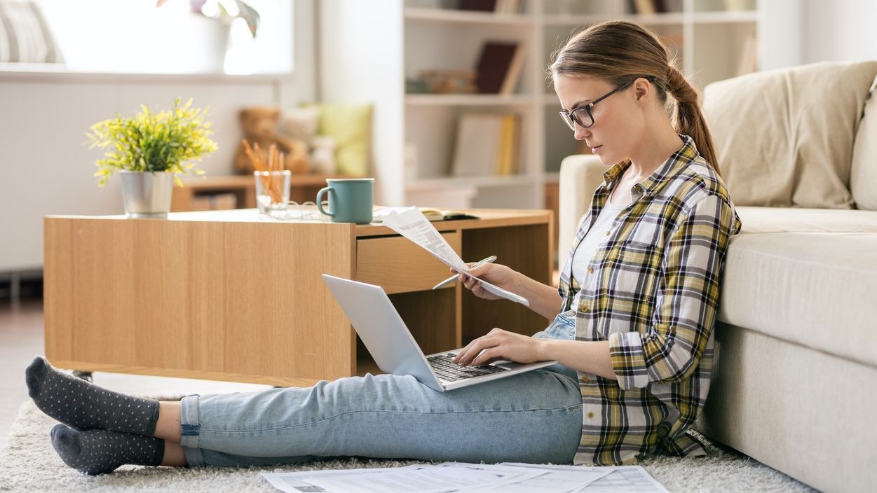 Junge Frau mit Brille sitzt auf dem Boden mit Laptop, Stift und einem Zettel.