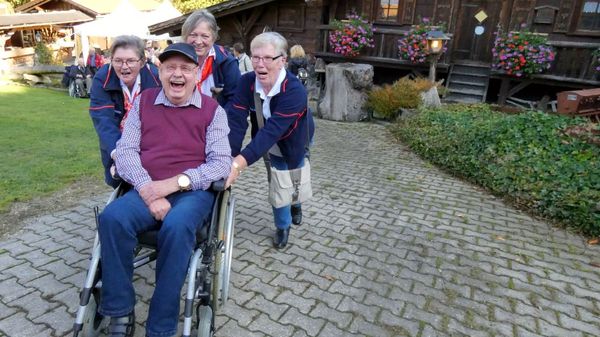 Lachender, älterer Rollstuhlfahrer wird von drei Frauen angeschoben.