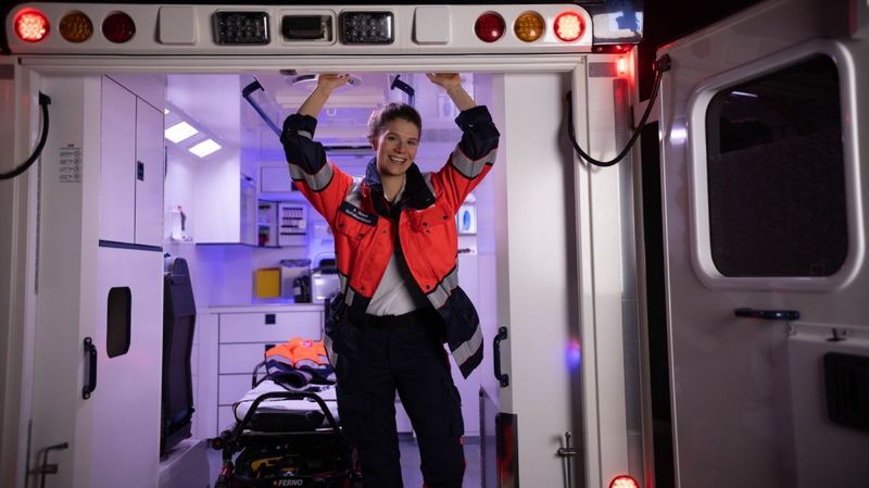 Eine junge, lächelnde Notfallsanitäterin im Heck eines Rettungswagens