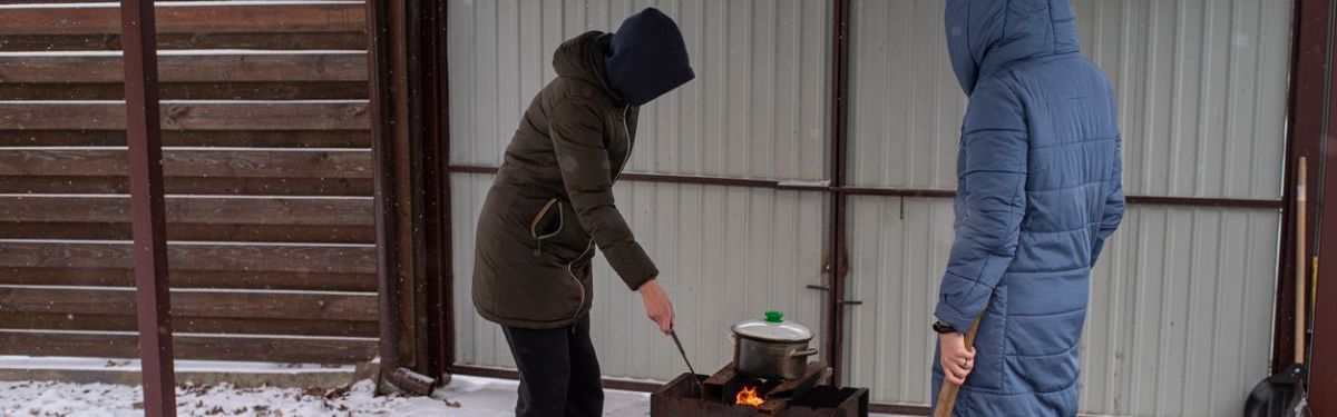 Zwei Menschen in Winterjacken kochen draußen etwas in einem Topf, der auf einem Grill steht.