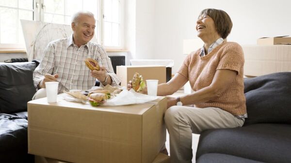 Ein älteres Paar isst lachend Sandwiches von gestapelten Umzugskartons.