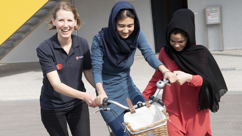 Eine lächelnde Frau bringt zwei jungen geflüchteten Frauen das Fahrradfahren bei. 