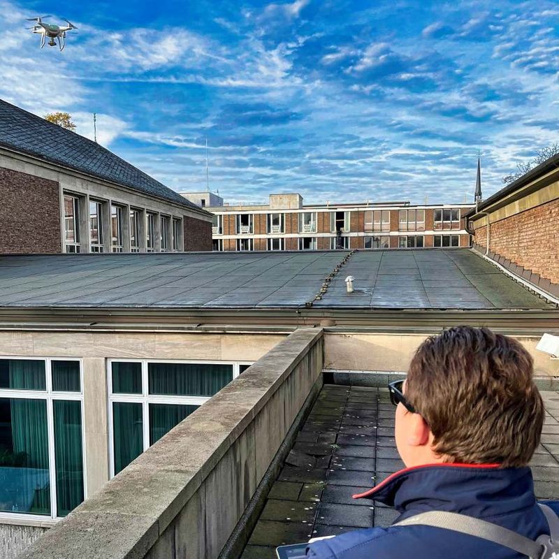 Mann mit kurzen braunen Haaren und blauer Jacke steuert eine Drohne über einem Gebäudekomplex. 