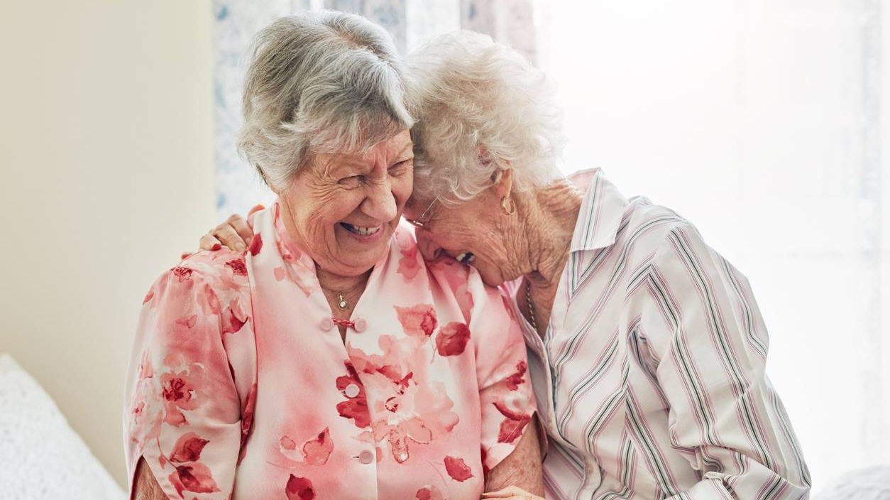 Zwei ältere Damen lachen herzlich und liegen sich dabei in den Armen.